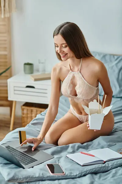 Jovem alegre em lingerie segurando caixa de macarrão e pagando com cartão de crédito na internet — Fotografia de Stock
