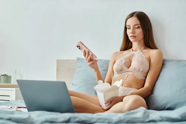 Вдумлива молода жінка в бежевій білизні тримає телефон і коробку з локшиною і дивиться на її ноутбук — стокове фото
