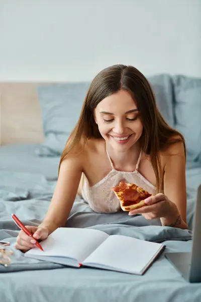 Atractiva mujer alegre en lencería acostada en la cama disfrutando de la pizza y tomando notas mientras se trabaja duro - foto de stock