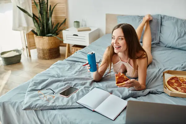 Atractiva mujer alegre acostada en la cama con pizza y refrescos y mirando hacia otro lado mientras trabaja en el ordenador portátil - foto de stock