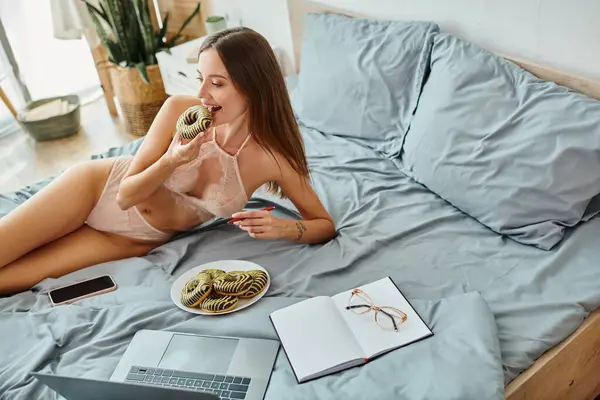 Alegre atraente mulher no sedutor lingerie deitado na cama e comer donut enquanto trabalhava em casa — Fotografia de Stock