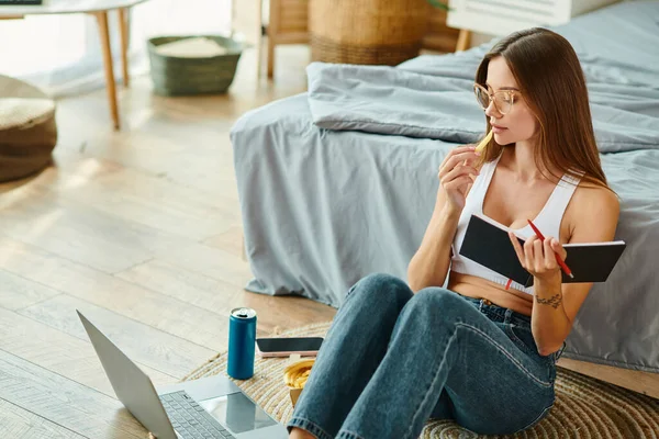 Pensive attractive woman with glasses sitting on floor while working remotely and eating fries — Stock Photo