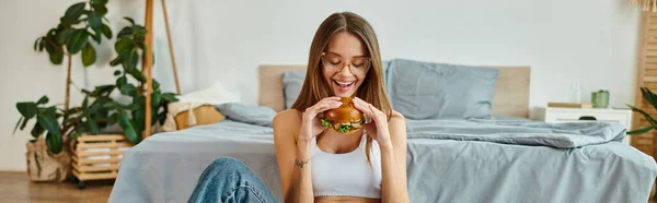 Mulher atraente alegre com óculos e cabelos longos desfrutando de delicioso hambúrguer enquanto em casa, banner — Fotografia de Stock