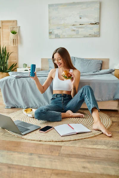 Mujer atractiva feliz en ropa casual sentado en el suelo y comer hamburguesa mientras trabaja en su computadora portátil - foto de stock