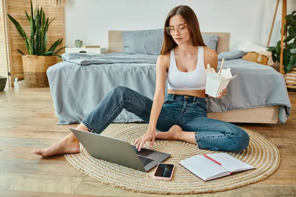 Mujer atractiva pensativa en ropa de casa acogedora sentada en el suelo con fideos y trabajando duro en el ordenador portátil - foto de stock