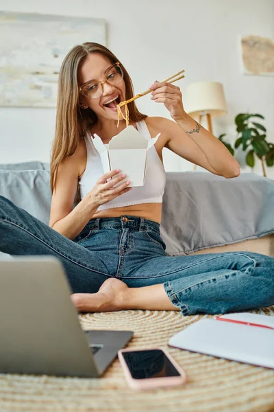 Alegre joven en ropa de casa con gafas y pelo largo disfrutando de fideos mientras trabaja de forma remota - foto de stock