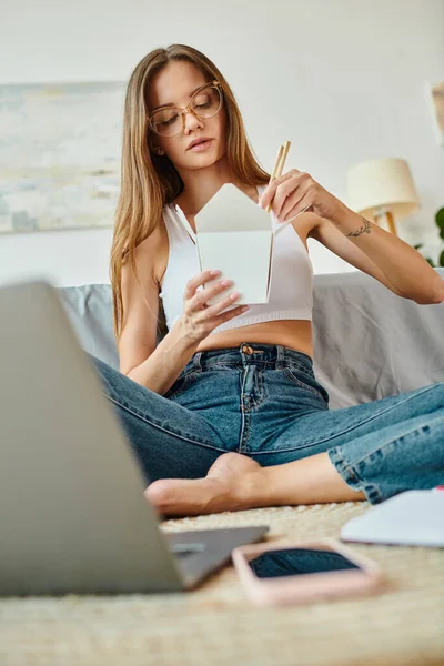 Привлекательная женщина с длинными волосами сидит на полу и ест лапшу во время работы на ноутбуке — стоковое фото
