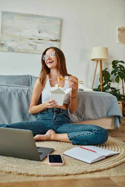 Mujer atractiva alegre con gafas en ropa de casa disfrutando de fideos mientras trabaja desde casa de forma remota - foto de stock