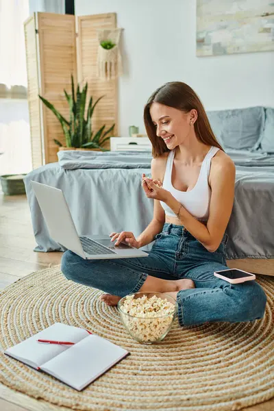Jolly beautiful woman sitting with her legs crossed and smiling at laptop with popcorn in hand — Stock Photo