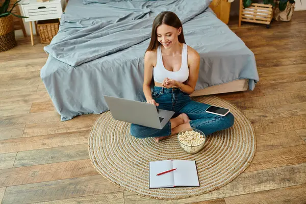 Joyeuse jeune femme en tenue de maison aux cheveux longs souriant à son ordinateur portable et mangeant du pop-corn frais — Photo de stock