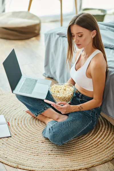 Appealing woman looking at her mobile phone while working at laptop and holding popcorn bowl — Stock Photo