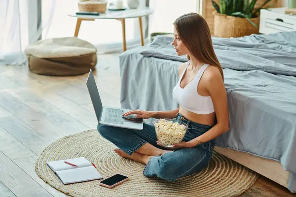 Mulher atraente em casa confortável sentado no chão e trabalhando em seu laptop com pipoca na mão — Fotografia de Stock
