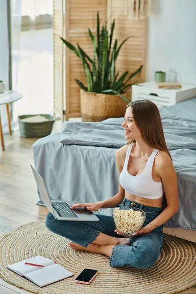 Alegre atractiva mujer en ropa de casa sentado en el suelo con palomitas de maíz mientras se trabaja de forma remota en el ordenador portátil - foto de stock