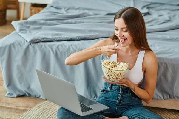 Atractiva mujer sentada en el suelo con las piernas cruzadas comiendo palomitas de maíz y viendo películas en el portátil - foto de stock
