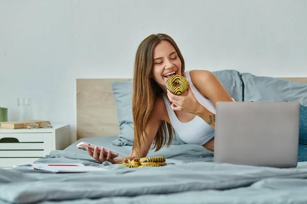 Joyous beautiful woman with long hair holding phone and enjoying donut while working from home — Stock Photo