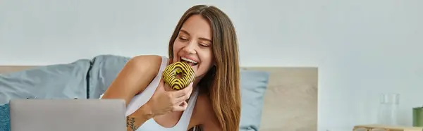 Jolly appealing woman with long hair enjoying donut while working at laptop at home, banner — Stock Photo