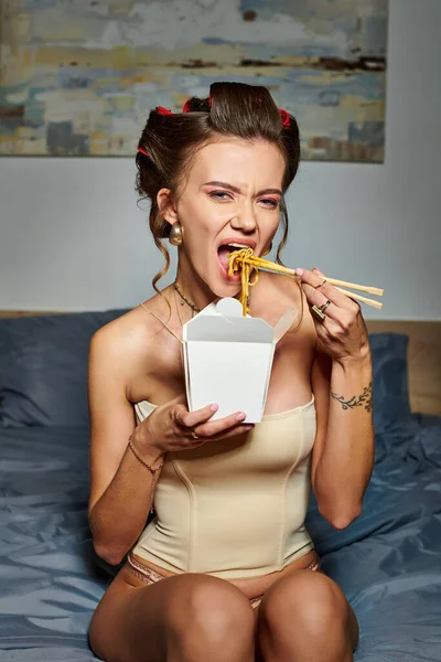 Sexy attrayant femme en beige corset avec des boucles de cheveux assis sur le lit et manger des nouilles asiatiques — Photo de stock