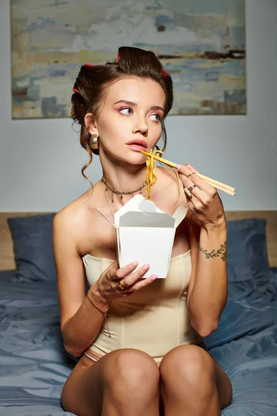 Appealing young woman in sexy beige corset with hair curlers eating delicious noodles on bed at home — Stock Photo