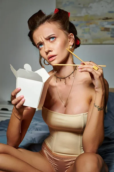 Confused woman in sexy beige corset with hair curlers sitting on bed with box of noodles in hand — Stock Photo