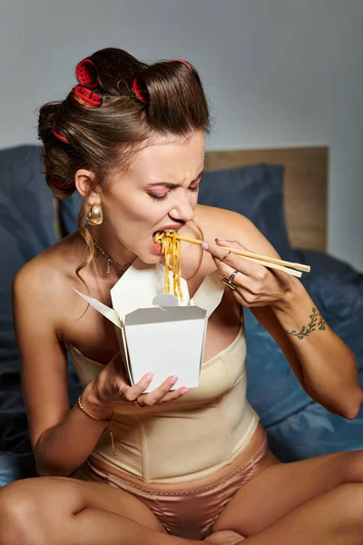 Atractiva mujer joven en corsé beige sexy comiendo fideos con palillos mientras está sentado en la cama - foto de stock