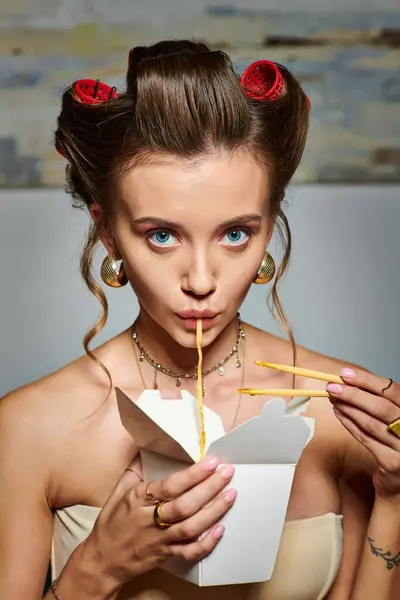 Hermosa mujer seductora con rizadores de pelo comiendo deliciosos fideos de caja usando palillos - foto de stock