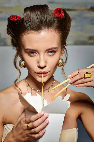 Attractive young woman with hair curlers eating tasty noodles from box and looking at camera — Stock Photo