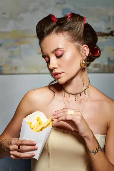 Alluring young woman with hair curlers in beige corset looking at pack of fries in her hands — Stock Photo
