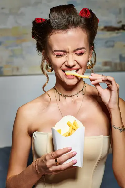 Jovem mulher emocional em espartilho bege com encrespadores de cabelo posando em casa e gostando de batatas fritas — Fotografia de Stock