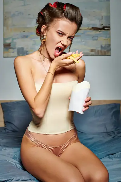 Attraente donna emotiva in corsetto sexy con bigodini capelli in posa con patatine fritte e guardando la fotocamera — Foto stock