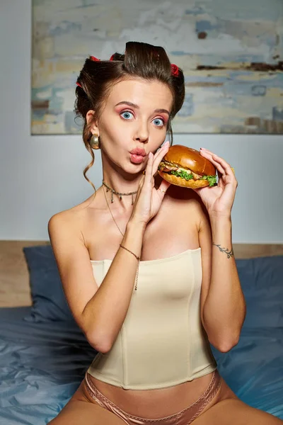 Alluring surprised woman with hair curlers and accessories holding burger and looking at camera — Stock Photo