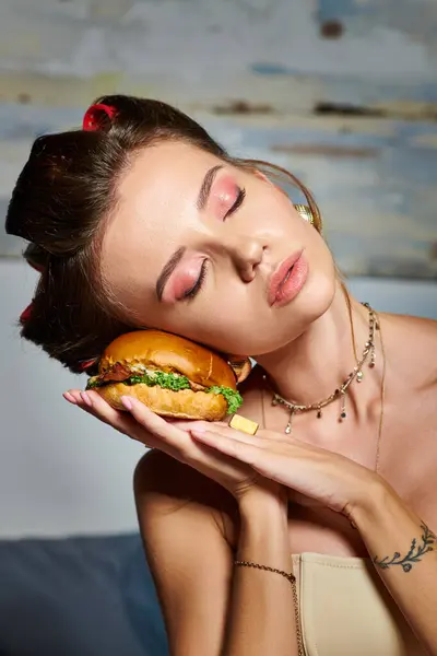 Attractive young woman with golden accessories and hair curlers putting her head on huge burger — Stock Photo