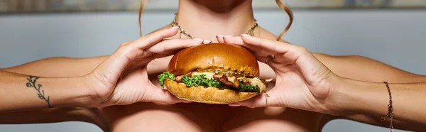 Обрезанный вид молодой женщины с аксессуарами и татуировкой на руке показывая огромный бургер, баннер — стоковое фото