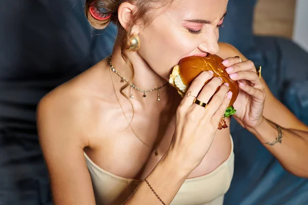 Alluring woman with hair curlers and accessories in beige corset eating delicious burger on camera — Stock Photo