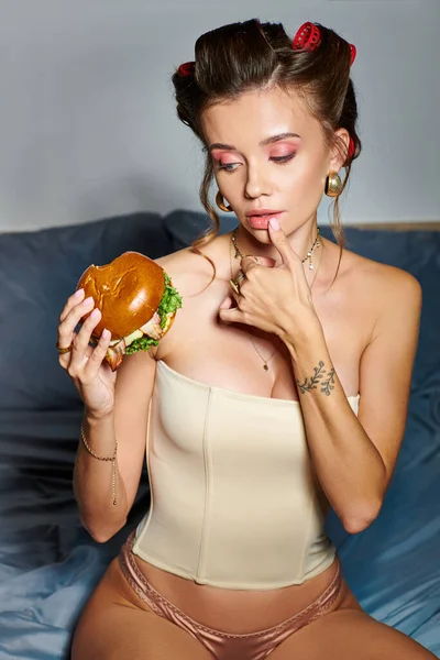 Séduisante jeune femme avec des bigoudis et accessoires appréciant le hamburger et la canette de boisson gazeuse — Photo de stock
