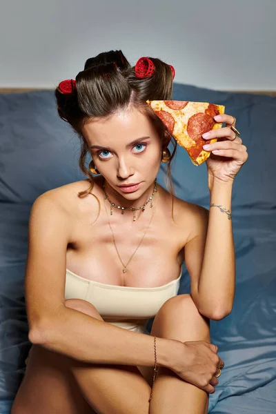Atractiva mujer con rizadores de pelo y accesorios posando con rebanada de pizza y mirando a la cámara - foto de stock