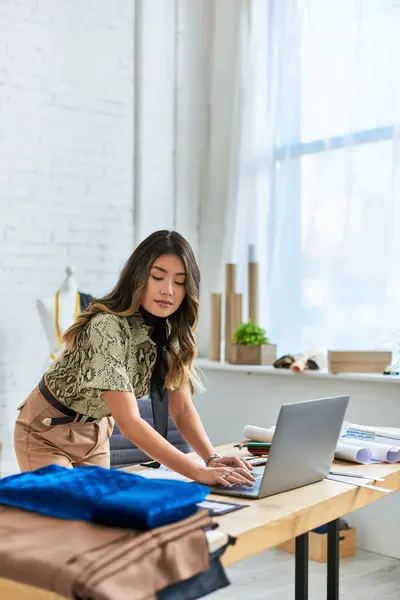 Trendy asian woman looking at fabric samples near laptop and sewing patterns in private atelier — Stock Photo