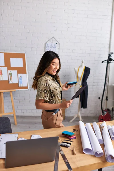 Inspirado estilista asiático mirando muestras de color cerca de la computadora portátil y patrones de costura en el estudio, moda - foto de stock