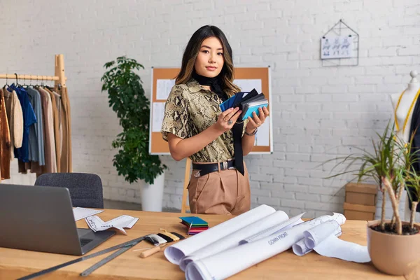 Attractive asian designer with color samples looking at camera near laptop and sewing patterns — Stock Photo