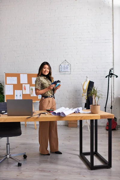 Designer asiático feliz com amostras de cor perto de laptop e padrões de costura no atelier de moda moderna — Fotografia de Stock