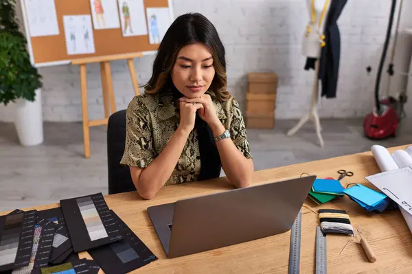 Reflexivo asiático diseñador de moda mirando portátil cerca de muestras de color en escritorio en atelier - foto de stock