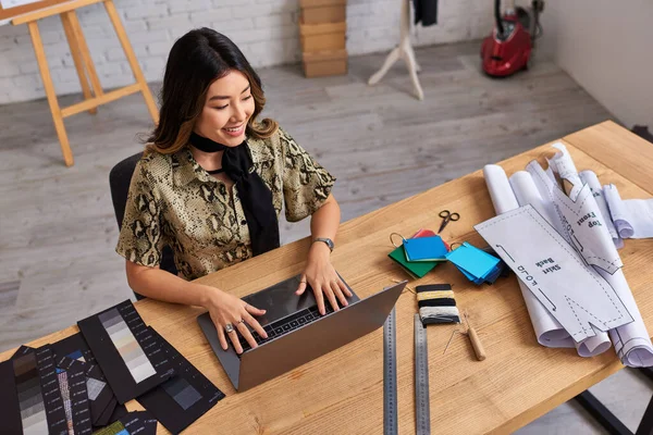 Alto ángulo vista de alegre asiático estilista mirando portátil cerca de color muestras en escritorio en atelier - foto de stock