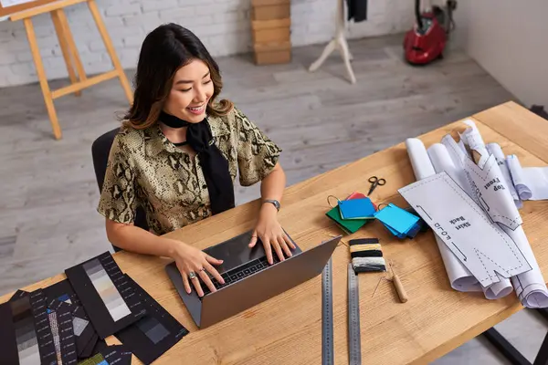 Alto ângulo vista de alegre asiático estilista olhando para laptop perto de amostras de cor na mesa no atelier — Fotografia de Stock