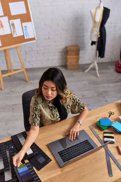 Designer de moda asiática criativa olhando para amostras de cor perto de laptop na mesa de trabalho, visão de alto ângulo — Fotografia de Stock