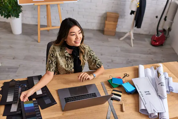 Щасливий азіатський стиліст моди посміхається біля ноутбука і кольорових годинників зі швейними візерунками в ательє — стокове фото