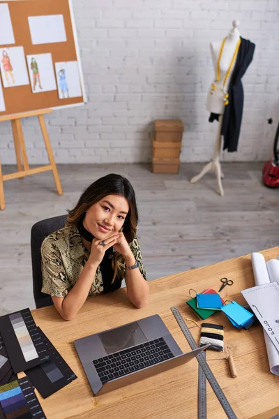 Alto ángulo vista de asiático estilista sonriendo en cámara cerca de portátil y muestras de color en estudio de moda - foto de stock