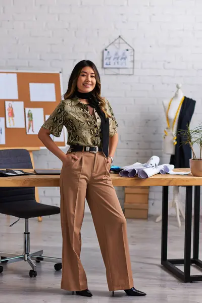 Fröhliche und trendige asiatische Designerin posiert mit den Händen in den Taschen am Schreibtisch im privaten Atelier — Stockfoto