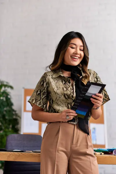 Pleased and trendy asian clothes designer looking at color swatches in private fashion workshop — Stock Photo