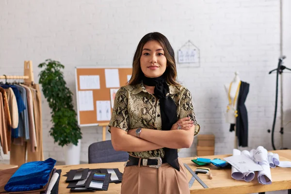 Successful happy asian clothes designer with folded arms looking at camera near work desk in studio — Stock Photo
