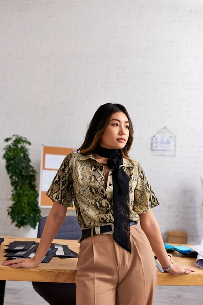 Reflexivo asiático ropa diseñador mirando lejos cerca trabajo escritorio en personal fashion studio, creatividad - foto de stock