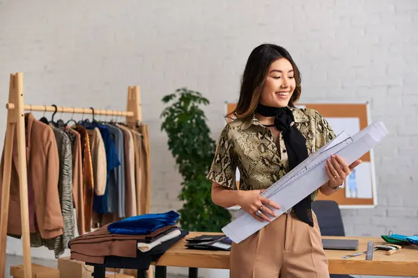 Young delighted asian designer standing with sewing patterns at work desk in modern personal atelier — Stock Photo